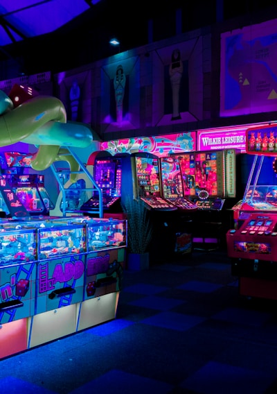 An indoor arcade filled with vibrant gaming machines lit with neon blue and pink lights. The scene includes various arcade games with flashing screens and colorful displays, and a large overhead structure resembling a green snake. The background shows illuminated signs and themed decor, contributing to an immersive entertainment atmosphere.