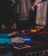 A person is engaged in a video game at a computer setup, using a keyboard and mouse. The scene is dimly lit, with the glow from the monitor reflecting off the hands of the player. Several other people are present in the background, indicating a social or competitive gaming environment.