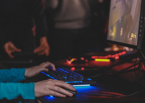 A person is engaged in a video game at a computer setup, using a keyboard and mouse. The scene is dimly lit, with the glow from the monitor reflecting off the hands of the player. Several other people are present in the background, indicating a social or competitive gaming environment.
