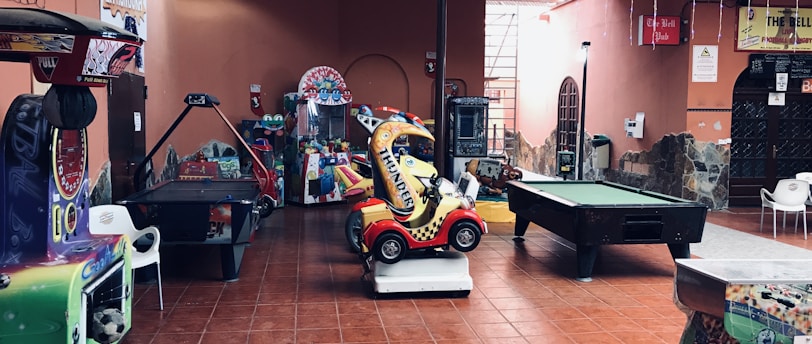 An indoor arcade area featuring various gaming machines, including an air hockey table and a ride-on toy car. The room has a tiled floor and several colorful gaming devices scattered around. Chairs and arcade signage are visible, creating a playful and entertainment-focused atmosphere.