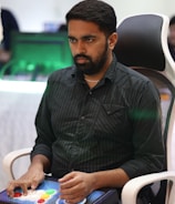 A person is seated in a chair, focused intently on something, with hands positioned on a gaming console or arcade controller. The setup appears to be indoors, with visible screens and lighting in the background, suggesting an electronic gaming environment.