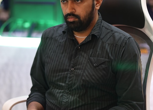 A person is seated in a chair, focused intently on something, with hands positioned on a gaming console or arcade controller. The setup appears to be indoors, with visible screens and lighting in the background, suggesting an electronic gaming environment.